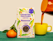 A box of Perennial coffee on a green ribbed surface and a cream background. A stemmed clementine sits to the left of the box and coffee is being poured into a yellow mug on the right side of the box.